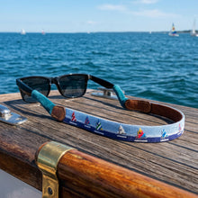 Load image into Gallery viewer, Needlepoint sunglass strap featuring sailboat motifs resting on a wooden boat deck overlooking the ocean