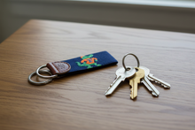 Load image into Gallery viewer, A navy blue woven key fob featuring a green and orange alligator design with an 'E' on its back, attached to a brown leather tab stamped with 's&amp;lE' and two silver metal key rings, shown on a plain white background.