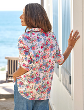 Load image into Gallery viewer, Woman wearing a floral button-up shirt with rolled sleeves, paired with wide-leg jeans, standing near the ocean.
