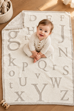 Load image into Gallery viewer, Smiling baby sitting on a soft, plush alphabet letter blanket on a wooden floor