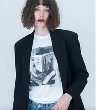 Load image into Gallery viewer, Model wearing a graphic tee featuring a person reading a newspaper, styled with blazer and jeans