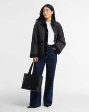 Load image into Gallery viewer, Model wearing quilted black jacket with gold buttons, paired with dark wide-leg jeans and tote bag