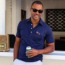 Load image into Gallery viewer, Man wearing navy Auburn polo shirt with chest pocket logo, holding beverage can outdoors