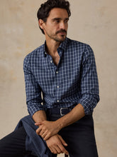 Load image into Gallery viewer, Male model seated wearing a navy plaid button-down shirt with sleeves rolled up