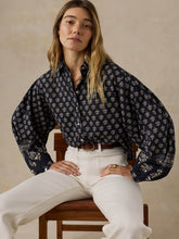 Load image into Gallery viewer, Model seated on wooden chair wearing oversized navy floral button-down blouse with white trousers