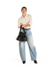 Load image into Gallery viewer, Model wearing wide-leg jeans holds Hammitt Gianna black leather drawstring bucket bag crossbody
