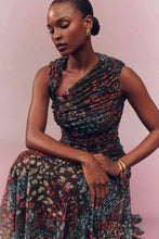 Load image into Gallery viewer, Model seated wearing a ruched sleeveless floral midi dress with draped neckline and gold accessories