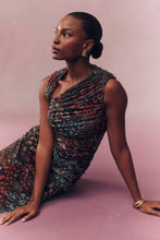 Load image into Gallery viewer, Model seated on pink backdrop wearing a draped sleeveless floral midi dress with gold accessories