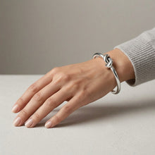 Load image into Gallery viewer, Model wearing a polished silver knot bangle bracelet on wrist resting on surface